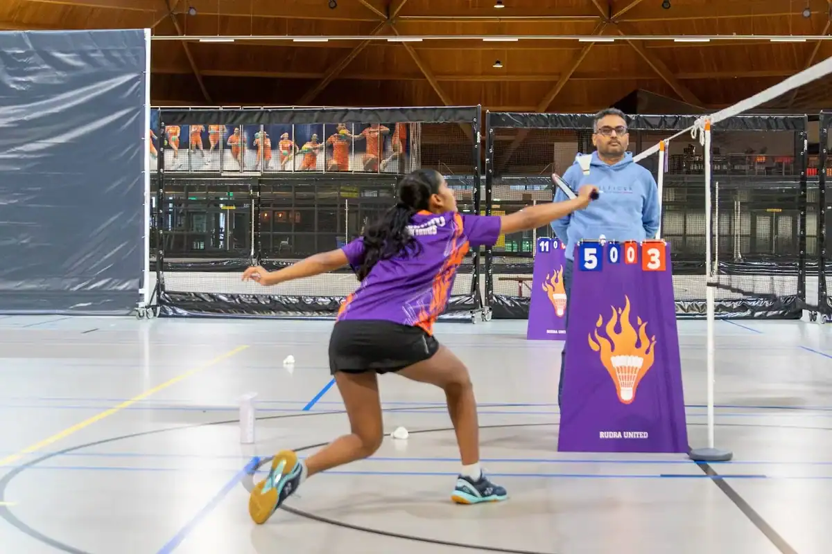 Junior player lunging for the shuttle next to the Rudra United banner during a youth league match in Eindhoven